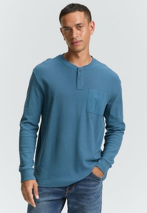 Langärmliges Henley-Shirt aus strukturiertem blauen Stoff. Mit einer Knopfleiste mit drei Knöpfen und einer Brusttasche. Trägt dazu eine dunkle Denim-Jeans.