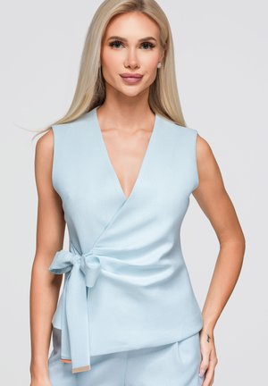 Femme blonde aux cheveux longs portant un haut cache-cœur sans manches bleu clair noué à la taille et un pantalon assorti, debout devant un fond uni.