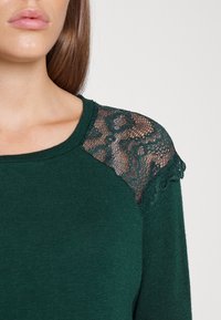Green sweater with a round neckline, featuring lace accents on the shoulder. The texture is soft, with a fitted design and delicate lace pattern.