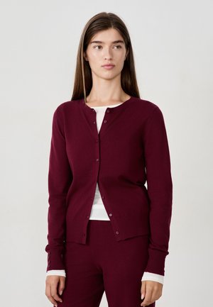Woman with long brown hair wearing a maroon buttoned cardigan and matching pants over a white long-sleeve shirt, standing against a plain background.