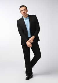 Mann im schwarzen Anzug und blauen Poloshirt steht mit gefalteten Händen und einem Bein übereinandergeschlagen vor einem schlichten hellen Hintergrund.