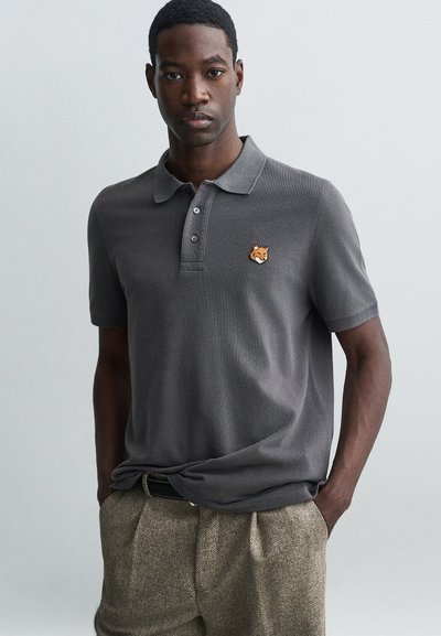 Polo gris à trois boutons, avec un logo brodé de tête de renard sur la poitrine gauche. Porté avec un pantalon beige texturé.