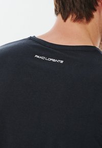 Czarny t-shirt z okrągłym dekoltem, z białym logo tekstowym "PAKO LORENTE" na tylnej części kołnierza. Miękka, gładka tekstura materiału.