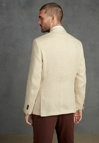 Blazer en lin beige clair avec une coupe ajustée, doté d'une fente à l'arrière et d'accents de boutons foncés. Porté sur un pantalon marron.