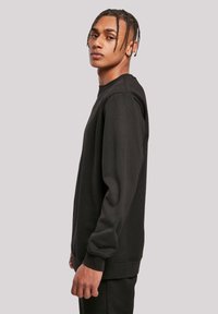 Junger Mann mit geflochtenem Haar, der ein schwarzes Langarmshirt trägt, seitlich vor einem schlichten hellgrauen Hintergrund steht.