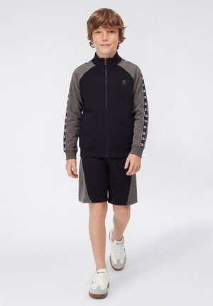 Junger Junge trägt eine schwarz-graue Jacke mit Reißverschluss und Shorts mit Logostreifen und geht in weißen Sneakern auf einem einfarbigen Hintergrund vorwärts.