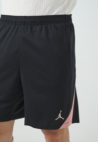 Svarta träningsshorts med en rosa sidopanel, meshstruktur och elastisk midja. Har en guld Jordan-logotyp längst ner till vänster.