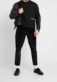 Sudadera negra con mangas en gris contrastante, acentuada con costuras verdes y un parche del logotipo. Combinada con pantalones cargo de pana negra y zapatos negros.