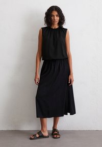 Vrouw met krullend haar, gekleed in een mouwloos zwart topje, een zwarte midi rok met een zijopening en zwarte sandalen met gespen, staande tegen een witte muur.