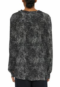 Personne aux cheveux tressés portant un chemisier à manches longues noir et blanc à motifs tachetés, vue de dos sur un fond blanc.