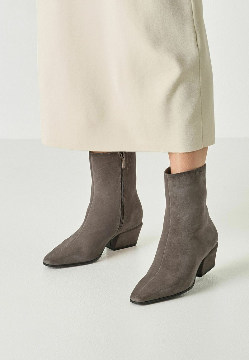 Femme portant des bottes mi-mollet en suède gris avec des talons blocs bas et des fermetures éclair latérales, associées à une jupe couleur crème.