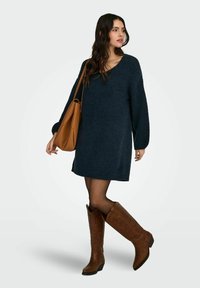 Granatowa dzianinowa sukienka z dekoltem w serek i długimi rękawami, połączona z intensywnie brązowymi kozakami za kolano z wytłoczonymi wzorami i obcasem typu słupek.
