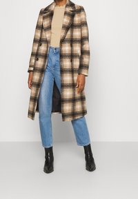 Gekonterter langer Mantel in Braun, Schwarz und Creme, aus strukturiertem Stoff, mit einem doppelt geknöpften Design und Reverskragen. Blaue Jeans und schwarze Stiefel.