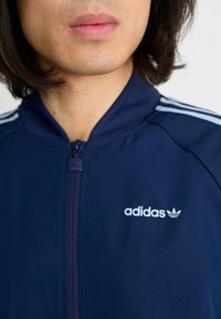 Tengerészeti kék, sima anyagból készült zipzáros dzseki, fehér Adidas logóval és három fehér csíkkal a vállakon.