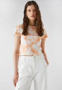 Korte mouwen, cropped t-shirt in een oranje en wit tie-dye patroon; voorzien van tekst "HET IS NOOIT TE LAAT VOOR PLEZIER" in het rood. Zachte stof.