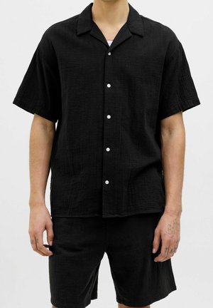 Uomo che indossa una camicia nera a maniche corte con bottoni e pantaloncini neri abbinati, in piedi davanti a uno sfondo semplice.