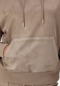 Sudadera de ante beige con un bolsillo tipo canguro, costuras y detalles dorados, cordones con puntas doradas y puños acanalados.