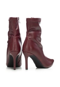 Pár bordó kožených kotníkových bot na vysokém podpatku s špičatými špičkami a štíhlými podpatky, zobrazených zezadu a z boku na bílém pozadí.