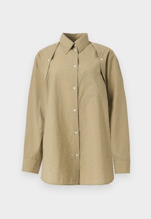 Femme debout portant une chemise oversize vert olive boutonnée avec des découpes aux épaules, un short noir longueur genou et des chaussures noires pointues.