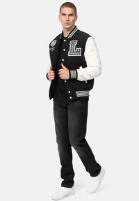 Chaqueta varsity en blanco y negro con parches bordados, puños y dobladillo acanalados, llevada sobre una camiseta negra, combinada con jeans negros y zapatillas blancas.