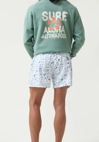 Sudadera verde con "SURF" y un gráfico floral en la parte posterior, combinada con pantalones cortos blancos que presentan patrones abstractos en azul y iconos.