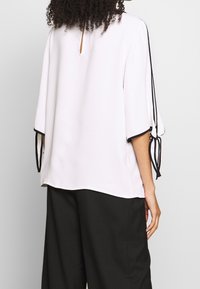 Blouse blanche avec des bordures noires et des manches amples, dotée de liens de chaque côté et d'une ouverture en forme de clef à l'arrière. Texture de tissu lisse.