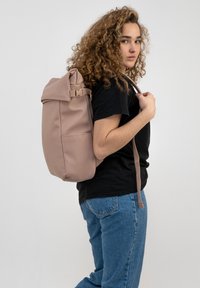 Zaino rosa morbido con design a roll-top, spallacci regolabili e finitura opaca, indossato su una spalla da una persona in abbigliamento casual.