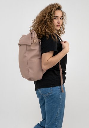 Un sac à dos rose doux avec un design à rabat, des bretelles ajustables et une finition mate, porté sur une épaule par une personne en tenue décontractée.