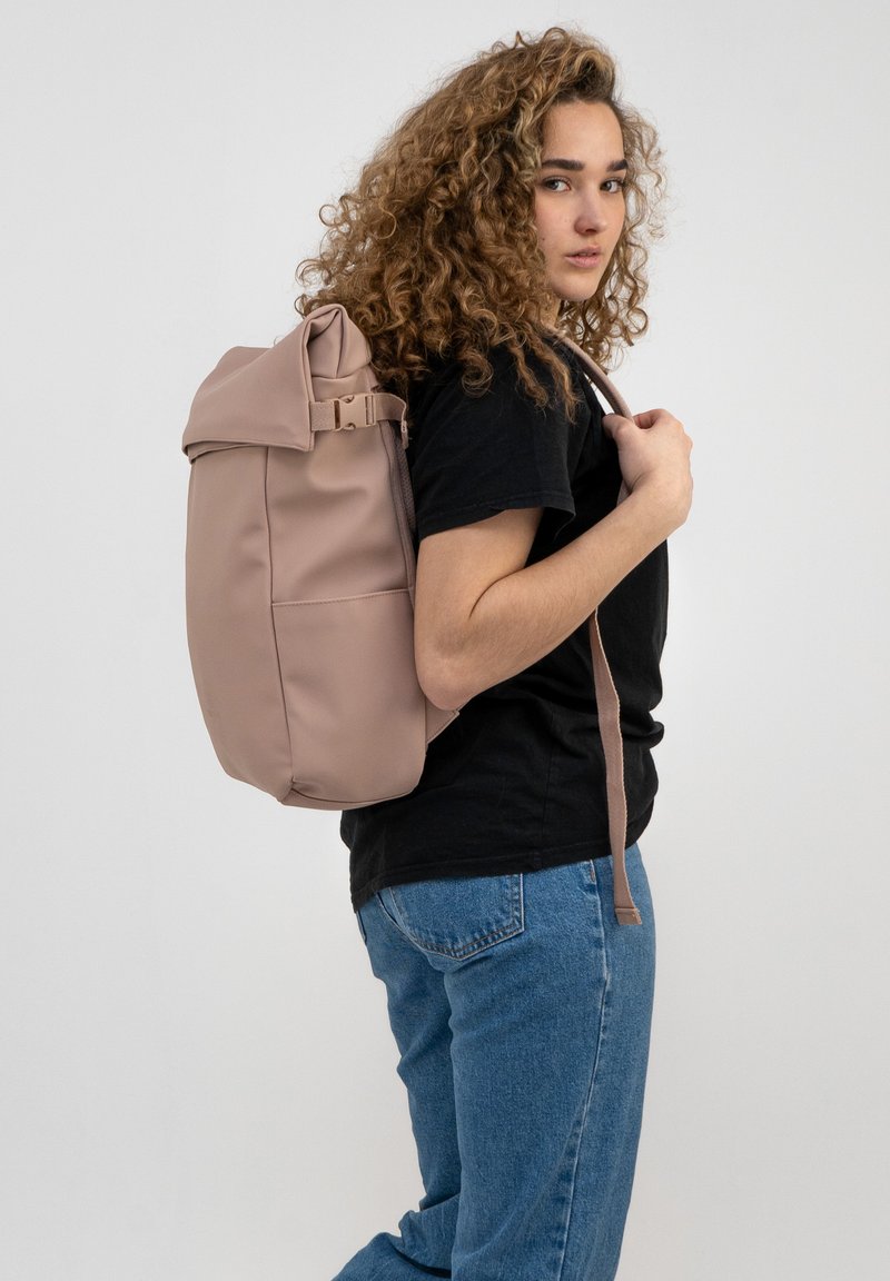 Zaino rosa morbido con design a roll-top, spallacci regolabili e finitura opaca, indossato su una spalla da una persona in abbigliamento casual.