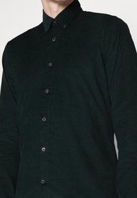 Tummanvihreä corduroy-paita, jossa on nappikaulus, etupaneeli nappikiinnityksellä ja teksturoitu kangaspinta. Slim fit -muotoilu.