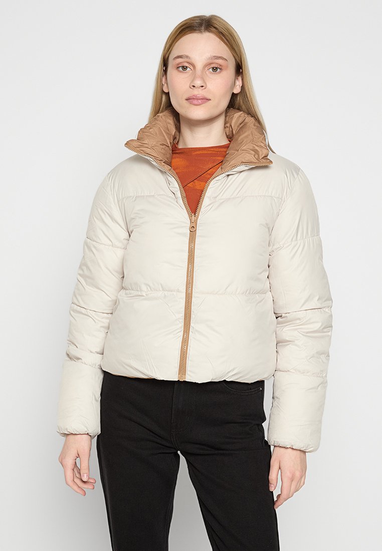 Chaqueta de plumas beige con un cuello marrón y cierre de cremallera. Presenta un patrón acolchado y puños de canalé, llevada sobre una blusa roja con estampado.
