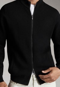 Cardigan noir à fermeture éclair avec col montant, poignets et ourlet côtelés. Tissu doux en maille avec poches latérales et fermeture éclair lisse.