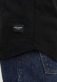 Fekete pamut póló, rajta a "JACK & JONES EST. 1990." felirattal ellátott címkével. A textúrált anyag és a ívelt hem a kék farmernadrág felett látható.