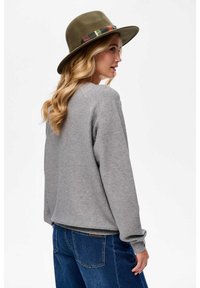 Kobieta z długimi blond włosami, nosząca oliwkową czapkę z wzorzystym paskiem, szary sweter i niebieskie dżinsy, stojąca bokiem na tle białym.