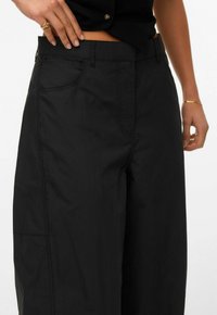 Osoba oblečená v černých kalhotách s vysokým pasem a černém krátkém topu, s jednou rukou opřenou poblíž pasu, nosící zlaté prsteny a náramek.