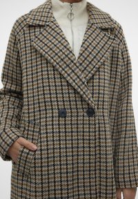 Smeđi i bež kaput s uzorkom houndstooth, dvostruko kopčanje, urezani ovratnik i dva crna dugmeta s bočnim džepovima.