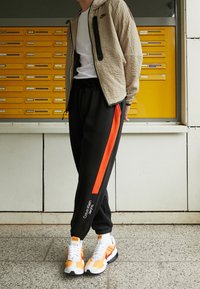 Beige textured jacket with black zipper, white t-shirt, black joggers featuring orange stripe and Calvin Klein logo, and orange/white sneakers.
