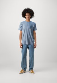 Lichtblauw t-shirt met korte mouwen met een grafische print op de borst, gecombineerd met losse lichtblauwe jeans en beige sneakers.