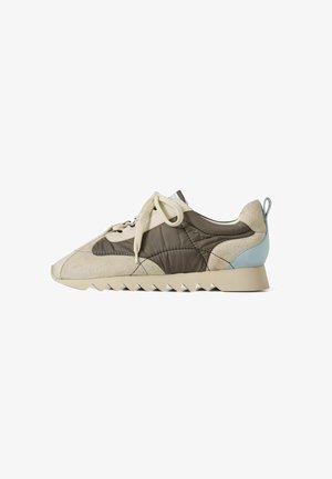 Zapatillas con parte superior de ante beige, paneles acolchados marrón oscuro, acento en el talón azul claro y suela de goma beige con bordes dentados. Diseño con cordones.