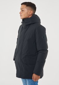 Giacca parka di un blu navy scuro con un colletto alto, grandi tasche frontali e una finitura lucida. Presenta un cappuccio removibile e un badge con logo sulla manica.