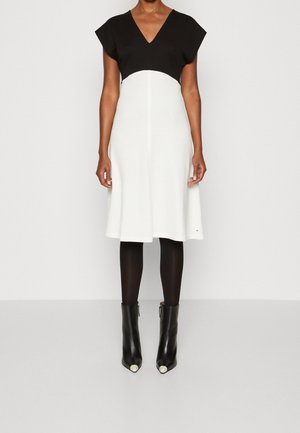 Vrouw draagt een zwart-witte jurk tot op de knie met V-hals, zwarte panty's en zwarte enkellaarzen met witte neus, staand op een witte vloer.