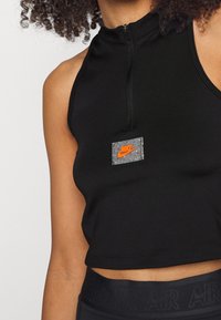 Kobieta w czarnym bezrękawnikowym topie z zamkiem błyskawicznym z przodu, z szarym naszywką z pomarańczowym logo Nike.