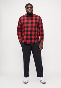 Camisa de manga larga a cuadros rojos y negros, abotonada, combinada con pantalones oscuros. Zapatillas deportivas blancas con detalles grises y logo azul.
