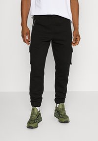 Svarta joggerbyxor i mjukt tyg, med sidofickor, dragkedjedetaljer och smalnande muddar. Matchas med gröna träningsskor.