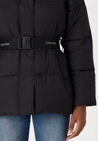 Black puffer jacket with a cinched waist belt featuring "Calvin Klein" branding. Soft, quilted texture with voluminous sleeves and pockets.