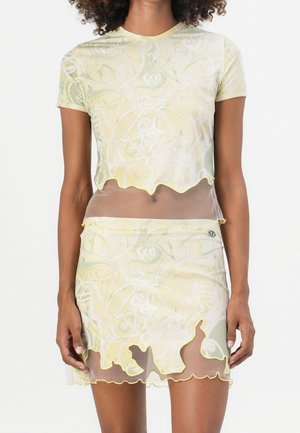 Top court transparent à manches courtes et mini-jupe assortie avec motif floral jaune et blanc et bords d'ourlet ondulés portés par une femme.