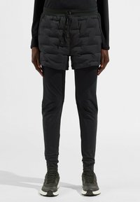 Shorts en duvet noirs par-dessus des leggings noirs ajustés, avec une taille à cordon de serrage, une texture matelassée et un tissu lisse et légèrement brillant sur les leggings.