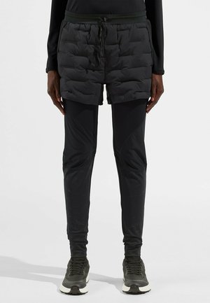 Schwarze Puffer-Shorts über schwarzen, enganliegenden Leggings, mit Kordelzugtaillierung, gepolsterter Textur und glattem, leicht glänzendem Stoff bei den Leggings.
