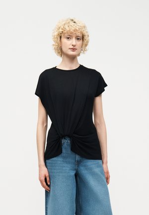 Ξανθιά γυναίκα με σγουρά μαλλιά, που φοράει μαύρο τοπ με κοντά μανίκια δεμένο στη μέση και μπλε τζιν με φαρδιά πόδια, που στέκεται μπροστά σε απλό φόντο.