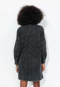 Robe noire à motifs avec des manches longues, présentant des motifs circulaires et en diamant. Fabriquée en tissu texturé, se terminant au-dessus du genou.